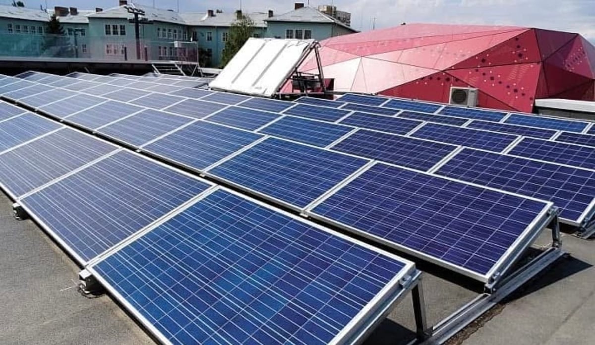 Array of blue solar panels installed on a rooftop with buildings and red structures in the background